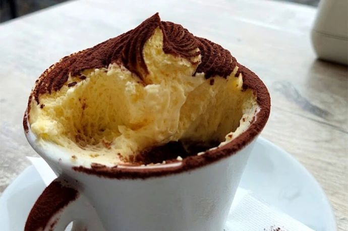 Close-up image of traditional Italian tiramisu served at Caffè Camerino. This classic dessert features layers of creamy mascarpone and coffee-soaked sponge, topped with fine cocoa powder, offering an authentic Roman café experience. Immagine ravvicinata del tradizionale tiramisù italiano servito da Caffè Camerino. Questo dessert classico presenta strati di cremoso mascarpone e savoiardi imbevuti di caffè, il tutto completato da una spolverata di cacao, per offrire un’autentica esperienza da bar romano.