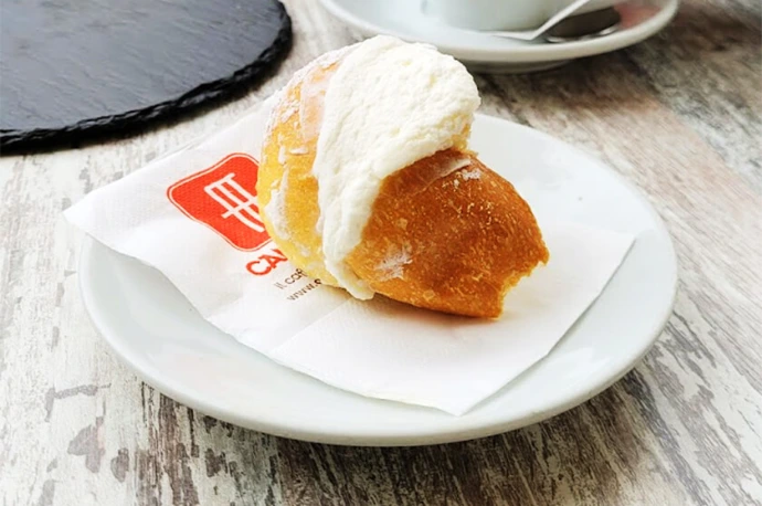 Close-up image of a traditional Roman maritozzo filled with fresh whipped cream, served at Caffè Camerino. This iconic Italian pastry represents the authentic flavors of Rome and pairs perfectly with a classic espresso for a true Roman café experience. Immagine ravvicinata di un tradizionale maritozzo romano farcito con panna fresca montata, servito da Caffè Camerino. Questo iconico dolce italiano rappresenta i sapori autentici di Roma e si abbina perfettamente a un espresso classico per una vera esperienza da bar romano.
