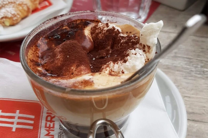 Close-up image of a traditional Italian espresso con panna served at Caffè Camerino. Featuring rich espresso topped with fresh whipped cream and a dusting of cocoa powder, this indulgent coffee reflects authentic Roman café tradition and premium Italian craftsmanship. Immagine ravvicinata di un tradizionale espresso con panna italiano servito da Caffè Camerino. Con un espresso ricco sormontato da fresca panna montata e una spolverata di cacao, questo caffè goloso riflette l’autentica tradizione del bar romano e la maestria italiana di qualità premium.